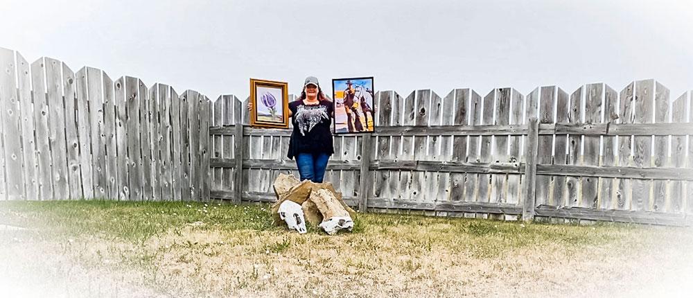 Woman standing in front of a gray fence holding two paintings